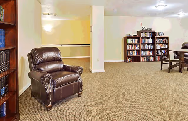 A spacious room with beige walls and carpeted floor featuring a dark brown leather armchair on the left, a wooden bookshelf filled with books in the background, and a wooden table with chairs on the right. The room is well-lit with ceiling lights and has a calm, inviting atmosphere.