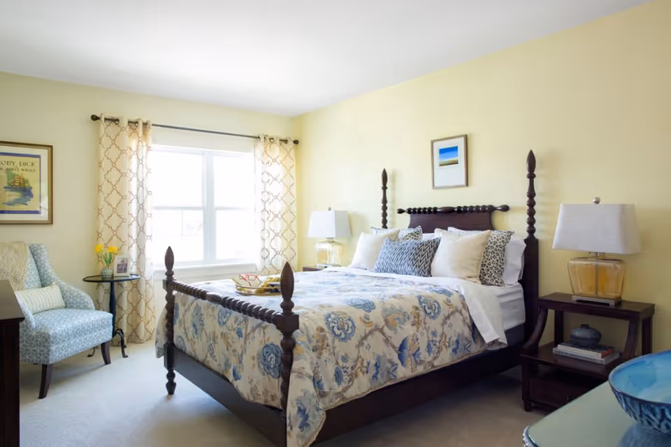 Sunlit bedroom with a wooden four-poster bed dressed in patterned bedding, bedside tables with lamps, an armchair by a window, and soft yellow walls.