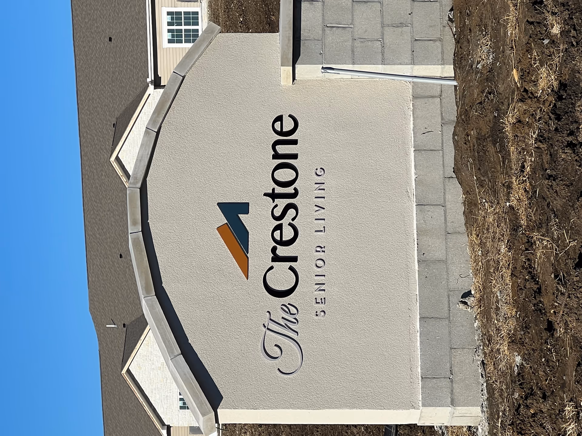 A large stone and stucco sign for The Crestone Senior Living facility with a logo above the text, set outdoors with a building and clear blue sky in the background.