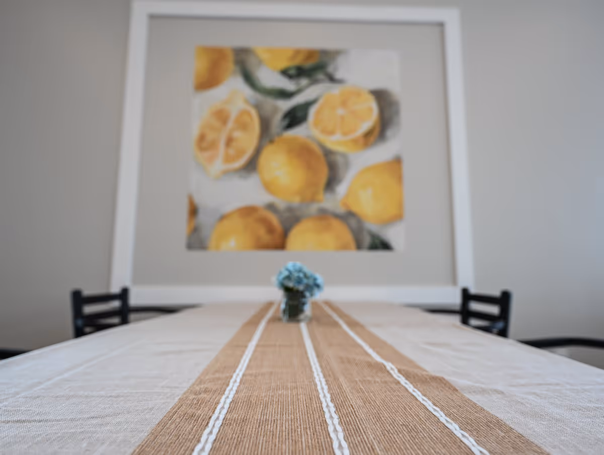A dining table with a striped runner and a small vase of blue flowers centered beneath a framed lemon painting.