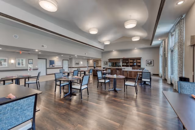 Spacious dining room with multiple tables and chairs arranged neatly on a dark wood floor. The room features large windows with curtains on the right side, modern ceiling lights, and a wooden cabinet with shelves against the far wall. The walls are painted light gray with framed artwork and sconces, creating a welcoming atmosphere.