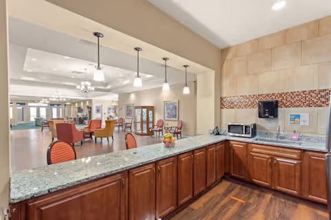 Interior view of a senior living facility showing a kitchen area with granite countertops, wooden cabinets, a microwave, and a sink. Beyond the kitchen is a spacious common area with multiple tables and chairs, some upholstered in red and orange, and decorative lighting fixtures hanging from the ceiling.