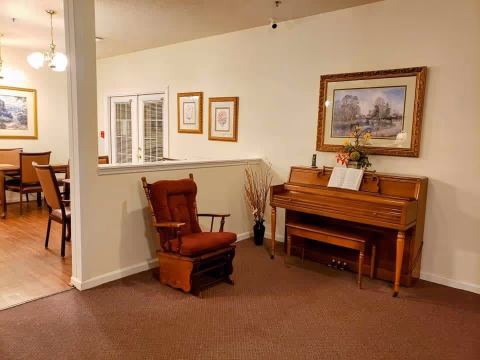 Interior view of a senior living facility showing a wooden piano with an open music book and a flower arrangement on top. Next to the piano is a cushioned wooden rocking chair. The walls are decorated with framed paintings and there is a partial view of a dining area with tables and chairs in the background.