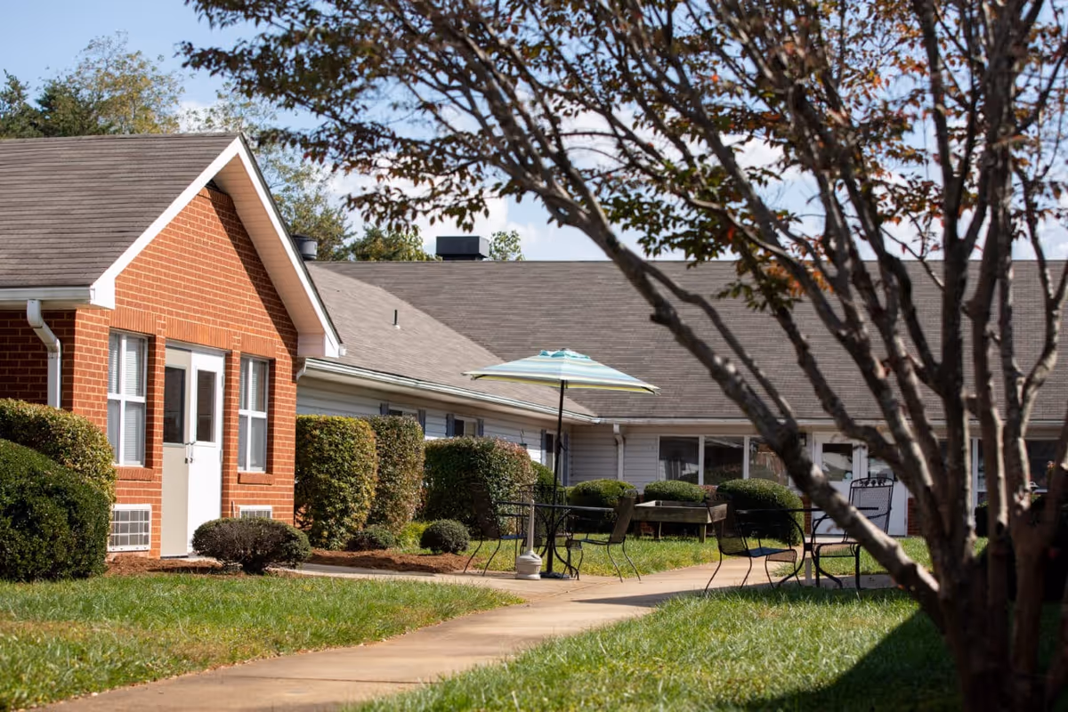 Outdoor view of a senior living facility with a brick building and a white building connected by a walkway. There are trimmed bushes along the buildings and a patio area with metal chairs and tables, one table with a blue and white striped umbrella. A tree with autumn leaves is in the foreground.