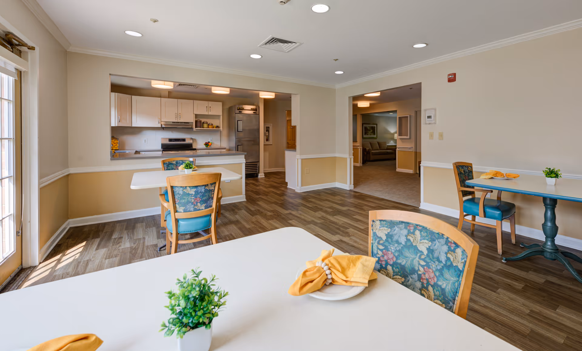A bright dining area with several tables and chairs featuring floral upholstery. The room has wood flooring, beige walls with white trim, and recessed lighting. In the background, there is a kitchen area with white cabinets, a stove, and a refrigerator. Small potted plants and folded yellow napkins are placed on the tables.