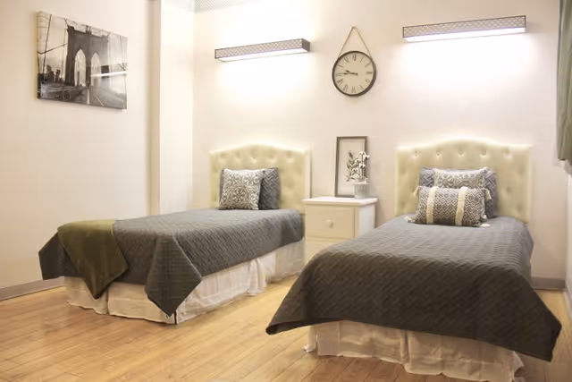 Two twin beds with tufted headboards and gray quilts flanking a nightstand under a wall clock in a well-lit bedroom.
