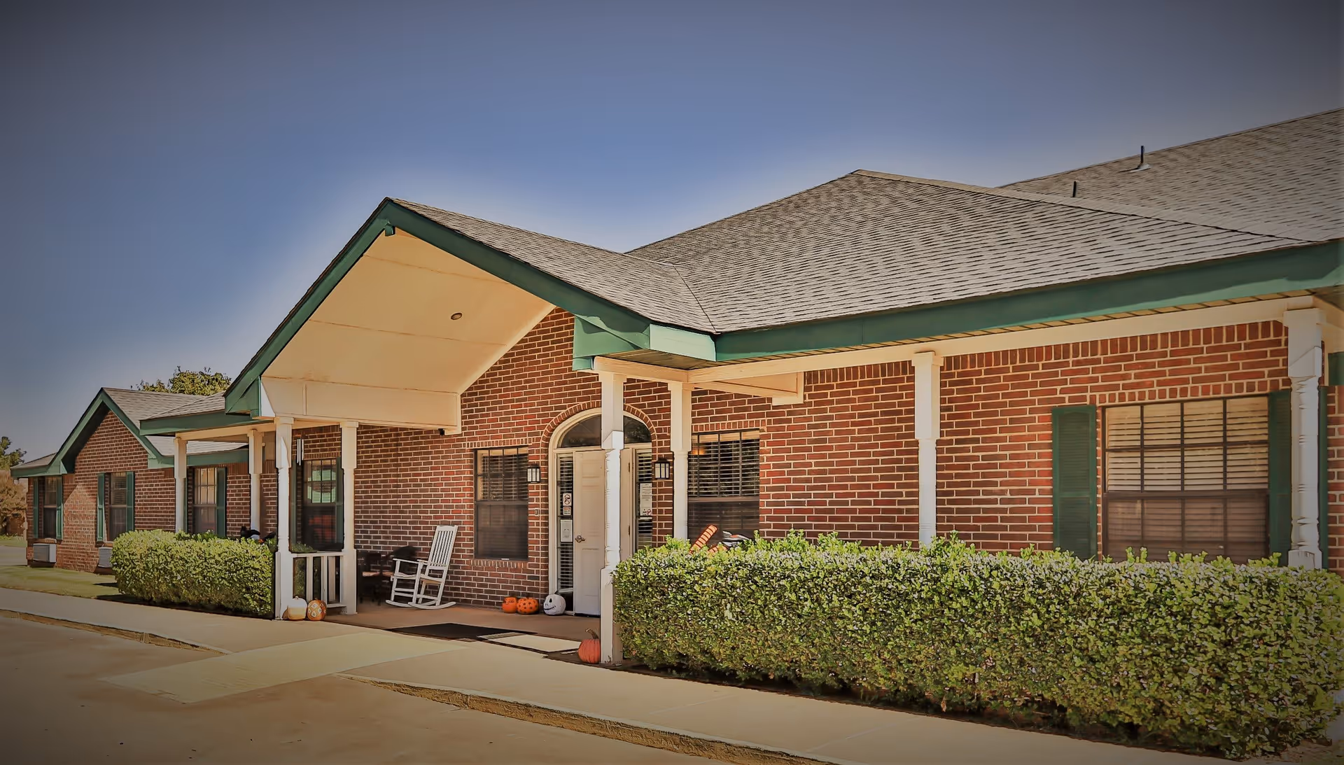 Exterior view of a single-story brick building with a covered entrance supported by white columns. The building has green trim and shutters, with a sidewalk and neatly trimmed bushes in front. There are rocking chairs and small pumpkins near the entrance.