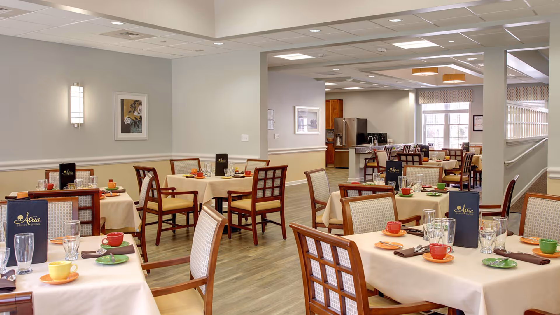 A dining room in a senior living facility with multiple tables covered in beige tablecloths, each set with colorful cups, saucers, plates, glasses, and silverware. Wooden chairs with cushioned seats surround the tables. The room has light-colored walls, framed artwork, and large windows allowing natural light to enter. A kitchen area with a refrigerator and countertop is visible in the background.