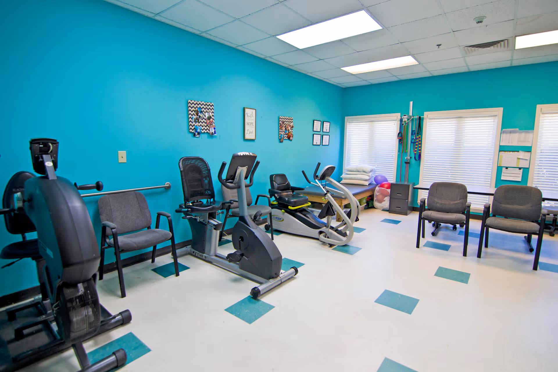 A brightly lit exercise room with turquoise walls featuring stationary exercise bikes, chairs, a therapy table with pillows, exercise balls, and resistance bands hanging on the wall. The floor has a white and teal checkered pattern, and there are windows with blinds letting in natural light.