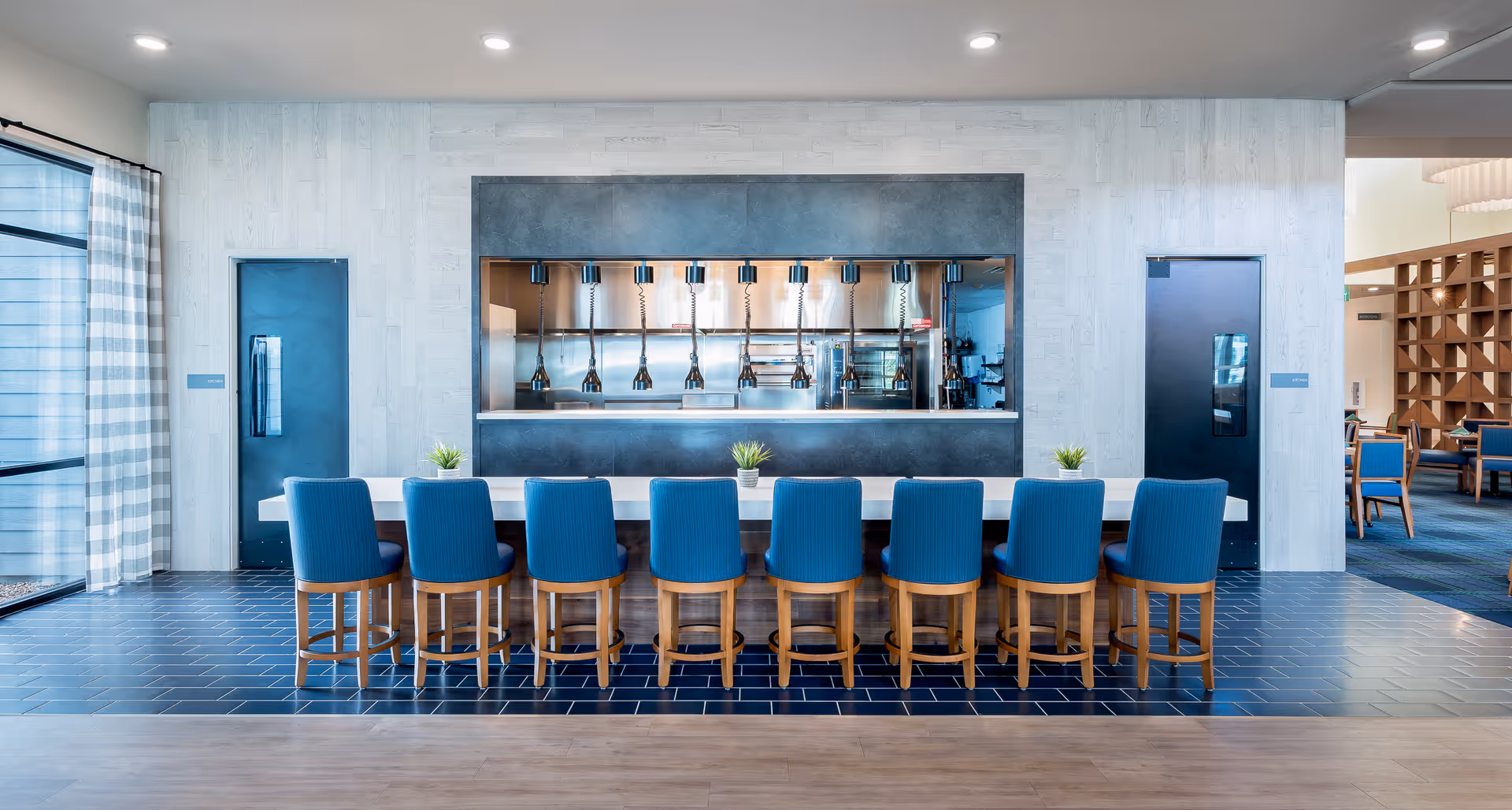A modern dining area with a long white table surrounded by ten blue upholstered chairs with wooden legs. Behind the table is a large pass-through window to a kitchen with hanging heat lamps. The floor near the table is covered with dark blue hexagonal tiles, transitioning to light wood flooring. Two black doors flank the kitchen window, and large windows with checkered curtains are on the left side, allowing natural light to enter.