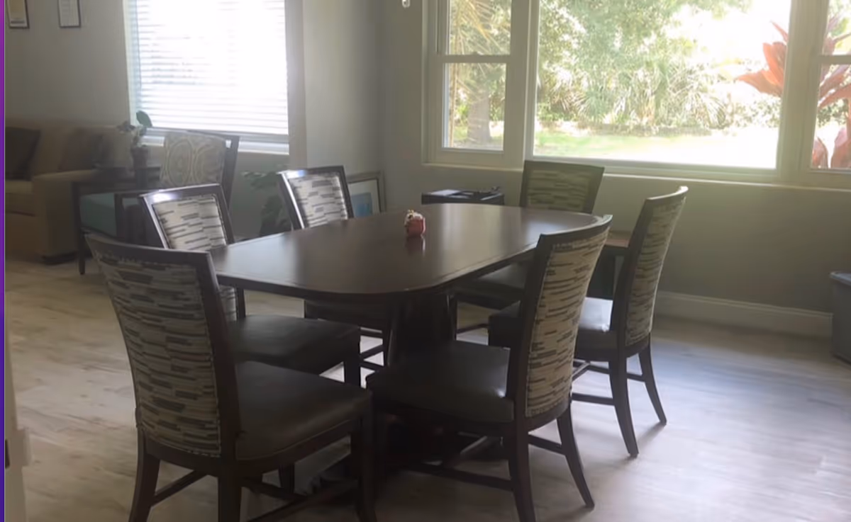 Dining room with a dark wooden table surrounded by six upholstered chairs and large windows letting in daylight.