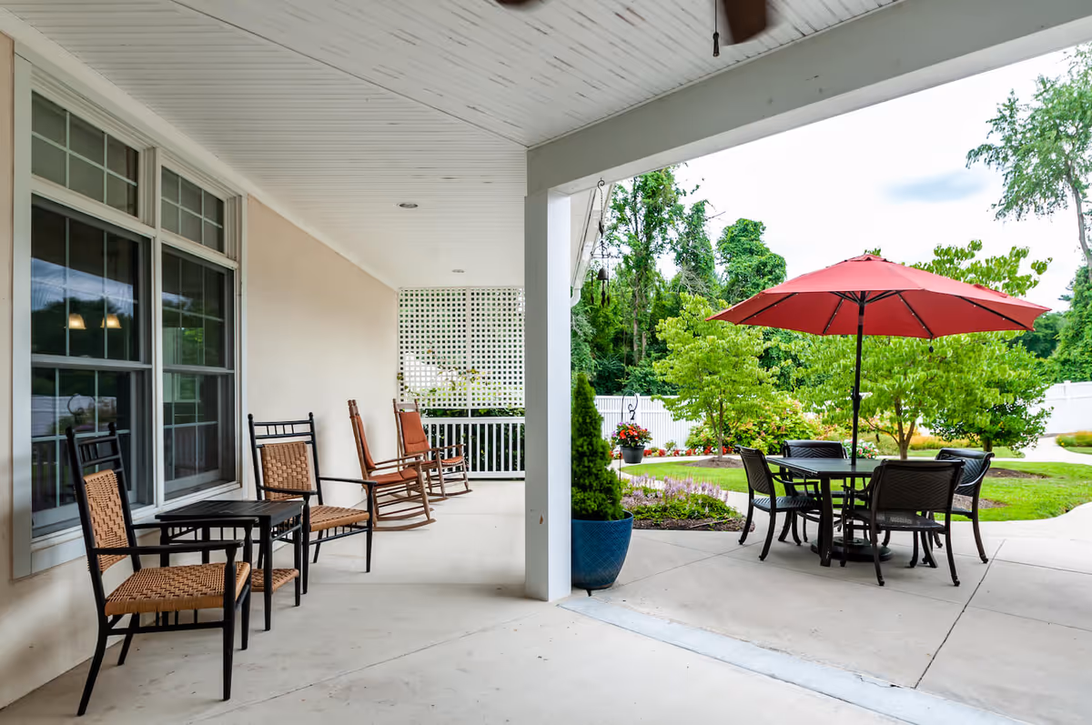 Covered patio with rocking chairs and wicker seating on the left and an outdoor dining table with a red umbrella overlooking a landscaped garden.
