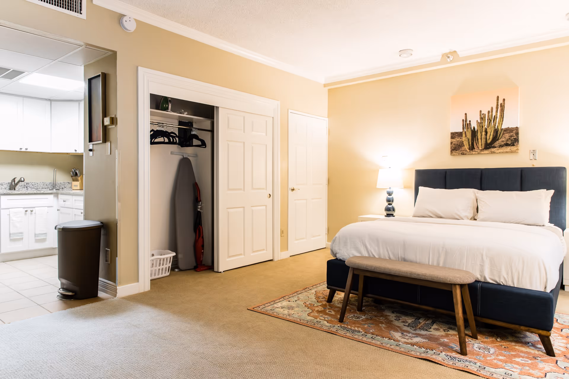 A well-lit bedroom with a large bed featuring white bedding and a dark blue upholstered headboard. A wooden bench sits at the foot of the bed on a patterned rug. To the left, there is an open closet with hangers, an ironing board, and a vacuum cleaner. A nightstand with a lamp is next to the bed, and a cactus-themed artwork hangs on the wall above the headboard. The room opens into a kitchen area with white cabinets and granite countertops.