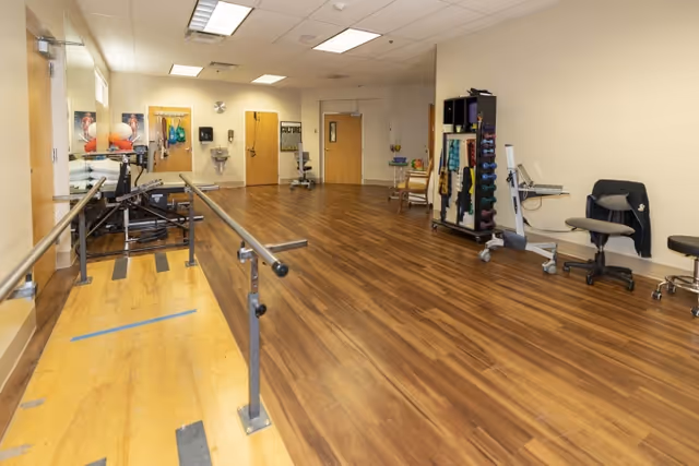 Spacious indoor therapy room with parallel bars, exercise equipment, and wood-look flooring.