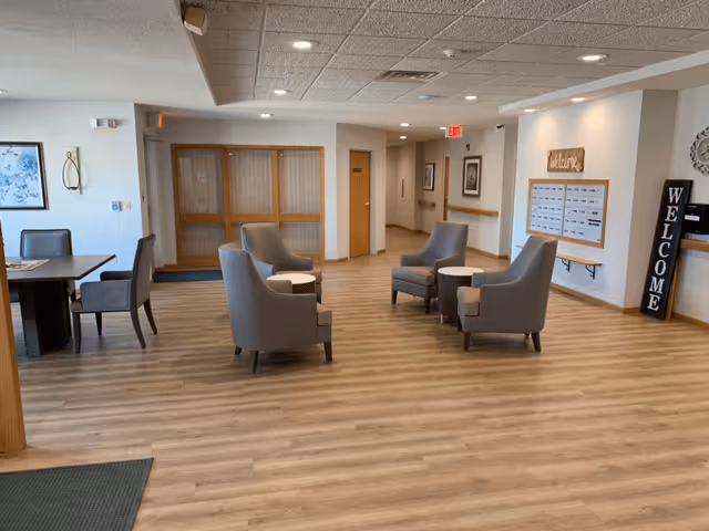 Spacious senior living lobby with gray armchairs arranged around small tables, mailboxes and a welcome sign along the wall.