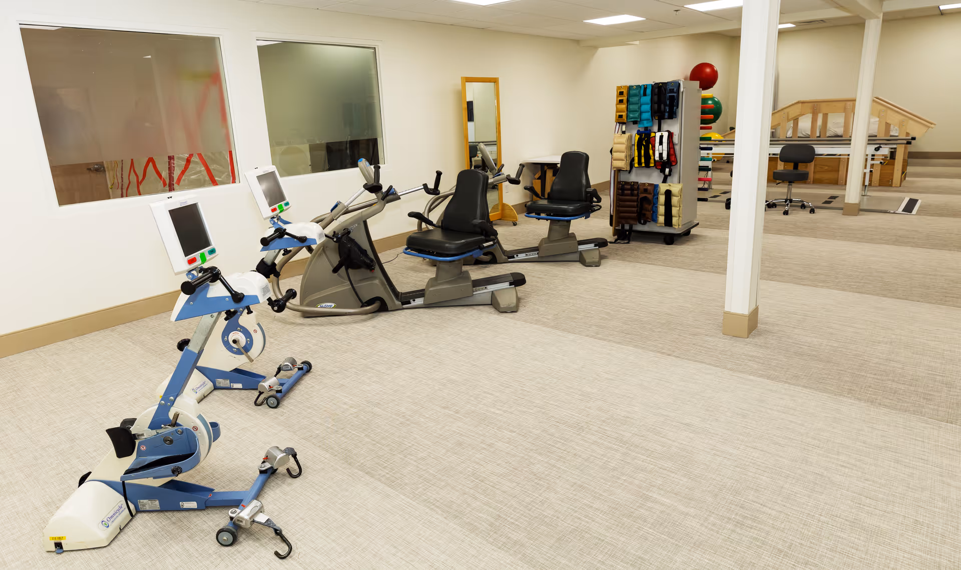 A spacious rehabilitation or physical therapy room with exercise bikes, recumbent bikes, a rack with various therapy bands and equipment, a mirror, and a wooden ramp with handrails in the background.