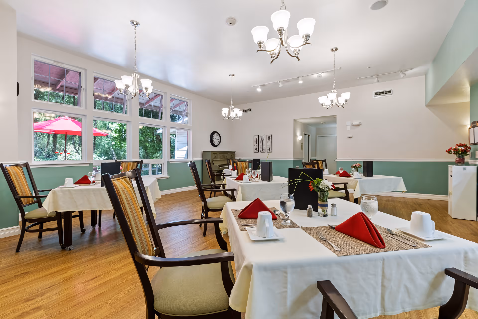 A bright dining room with several tables covered in white tablecloths, each set with red folded napkins, glasses of water, and silverware. The room has large windows showing greenery and a red patio umbrella outside. The walls are painted white and green, and there are multiple chandeliers hanging from the ceiling. Wooden chairs with striped cushions surround the tables.