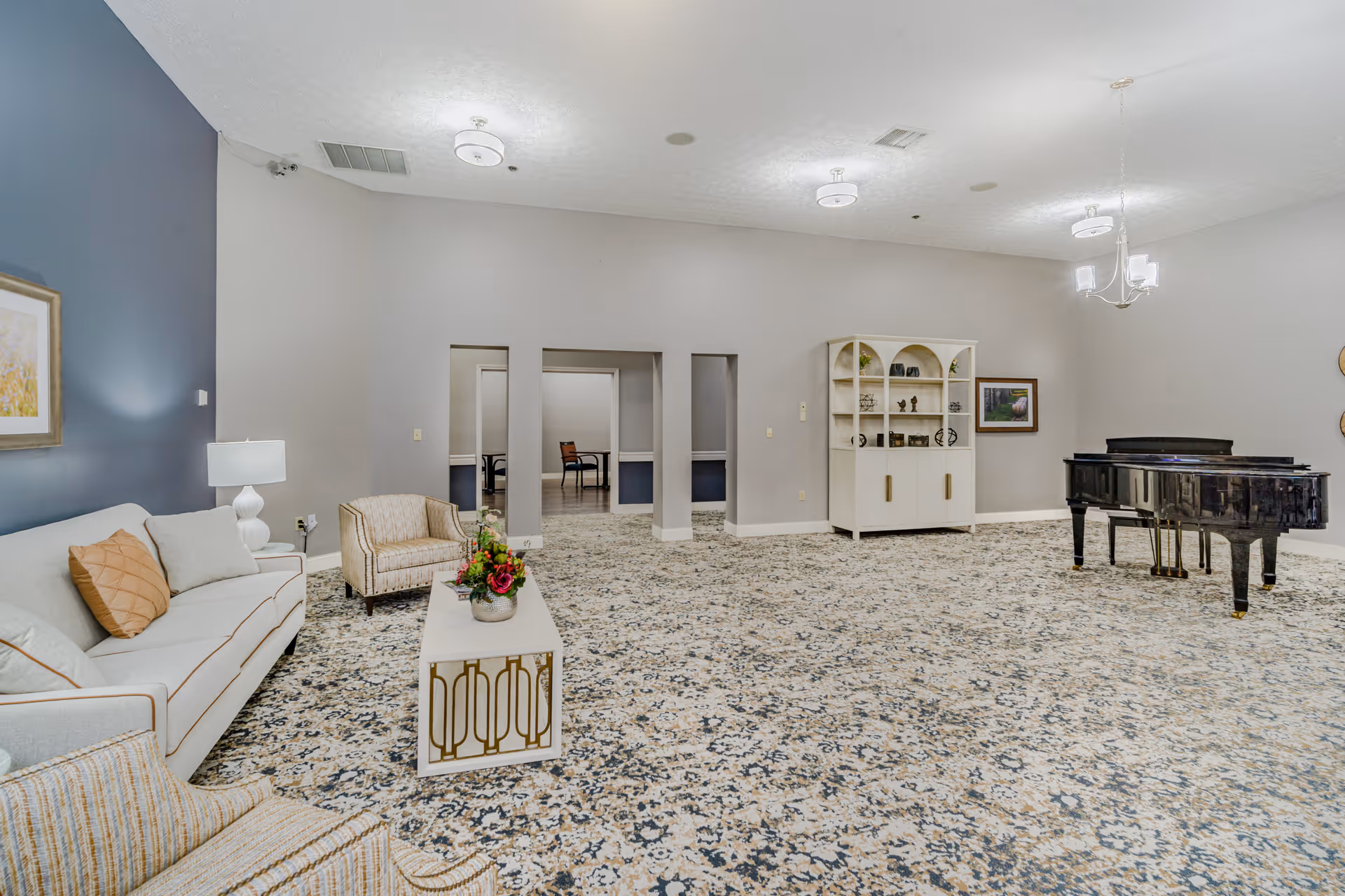 A spacious living room area with patterned carpet flooring, a white sofa with cushions, two armchairs, a white coffee table with a flower arrangement, a white cabinet with decorative items, a grand piano, and soft overhead lighting. The walls are painted light gray with one accent wall in blue, and there are three open doorways leading to other rooms.