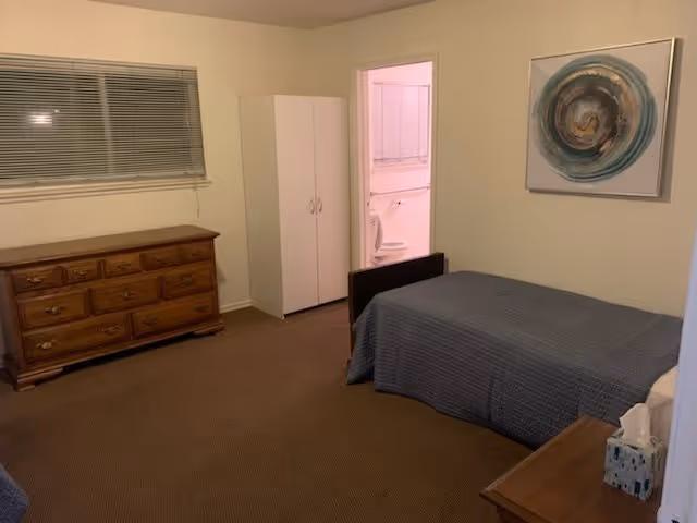 Simple furnished bedroom with a twin bed, wooden dressers, a wardrobe, wall art, and an open door to a bathroom.