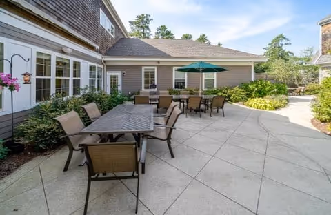 Sunlit outdoor patio courtyard with multiple tables and chairs, a green umbrella, and surrounding building and landscaping.