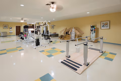 Bright rehabilitation/exercise room with parallel bars, weight machines, therapy stairs and patterned tiled floor.