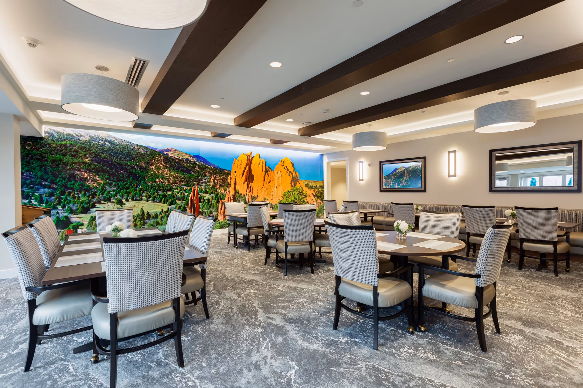 A spacious dining room with multiple tables and cushioned chairs arranged neatly. The room features a large wall mural of a scenic landscape with red rock formations and greenery. The ceiling has wooden beams and modern light fixtures, and the walls are decorated with framed pictures and mirrors.