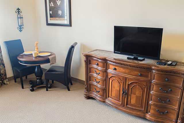 A small dining area with a round wooden table set for two with plates, cups, and a small decorative item. Two black chairs are positioned at the table. Next to the dining area is a wooden cabinet with ornate detailing, topped with a flat-screen TV and remote controls. A framed floral artwork hangs on the wall above the dining table.