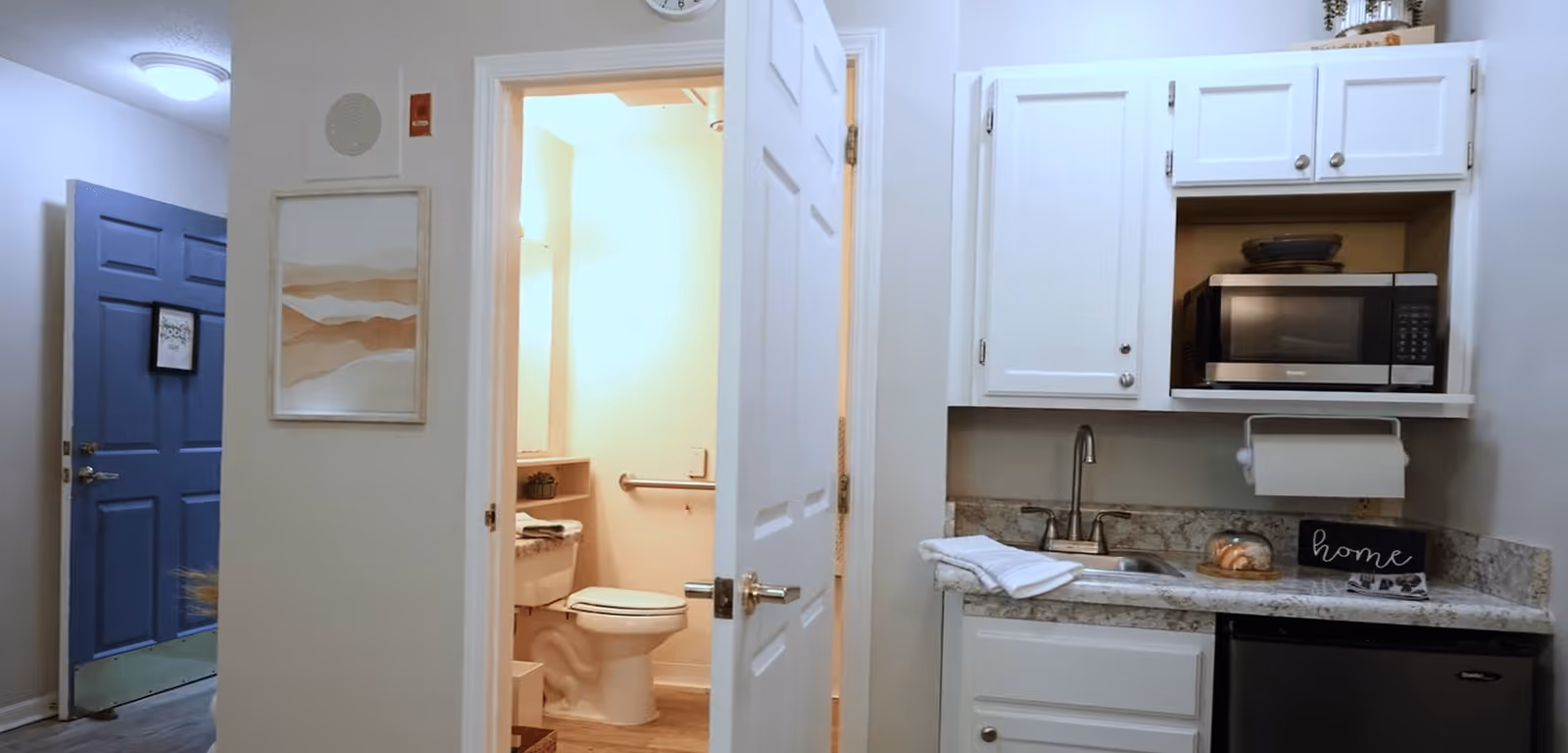 Interior view showing a small kitchenette area with white cabinets, a microwave, a sink, and a countertop with a towel and a decorative 'home' sign. To the left, an open door reveals a bathroom with a toilet and grab bars. A blue door is visible further left in the background.
