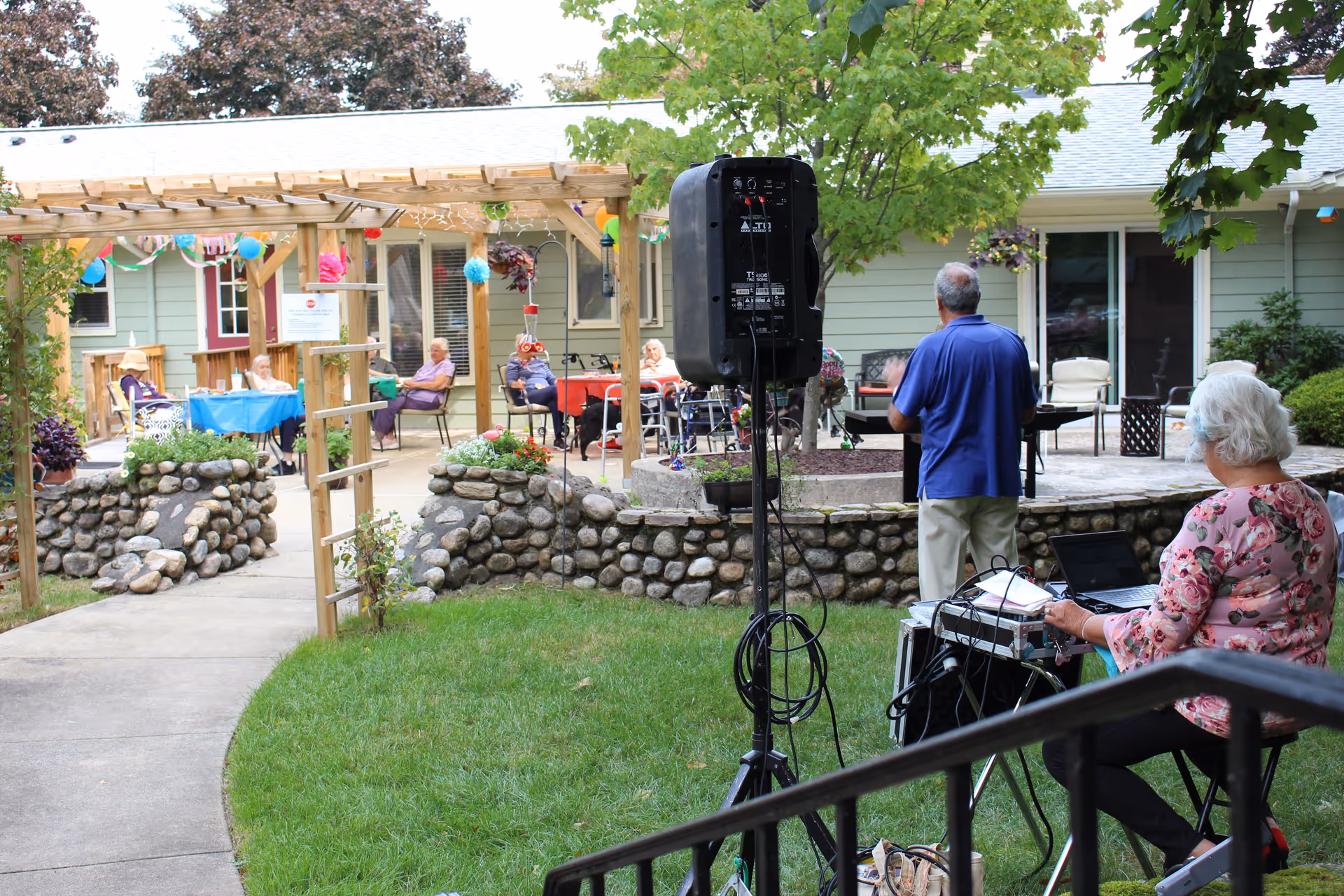 Outdoor courtyard area at Commonwealth House Assisted Living with elderly residents sitting at tables and chairs under a wooden pergola decorated with colorful balloons and streamers. A man in a blue shirt stands near a speaker, while a woman with white hair and a floral top operates audio equipment on a table. The scene is surrounded by greenery and stone walls.
