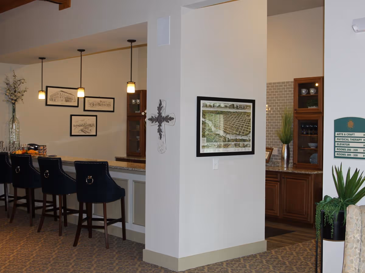 Interior view of a senior living facility showing a bar area with four dark blue high chairs, three pendant lights hanging above the bar, framed artwork on the walls, a decorative cross mounted on a white pillar, and a hallway with wooden cabinets and a plant in a vase.