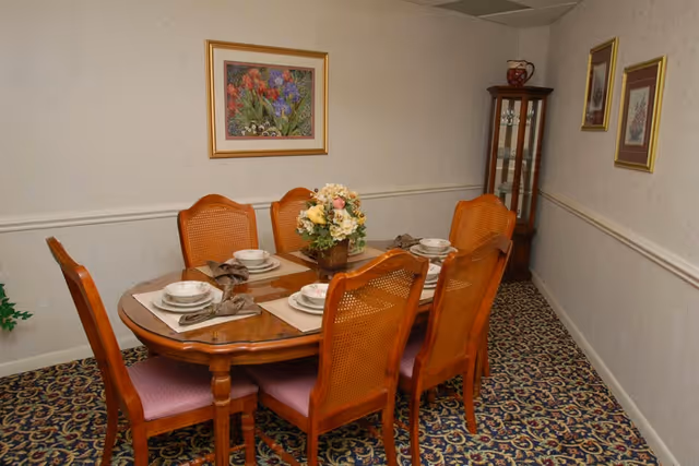 A dining room with a wooden oval table set for six people, each place setting includes a plate, bowl, and napkin. The chairs have wooden frames with woven backs and pink cushions. A floral centerpiece is on the table. The room has patterned carpet, light-colored wallpaper, and framed floral artwork on the walls. A glass-fronted wooden cabinet stands in the corner.