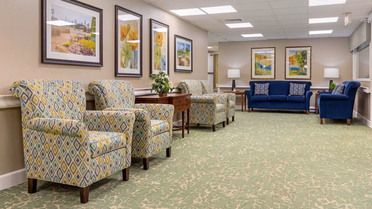 A spacious, well-lit common area with patterned armchairs and blue sofas arranged along the walls. The room features a green carpet with a subtle pattern, framed artwork on the walls, and table lamps providing additional lighting.