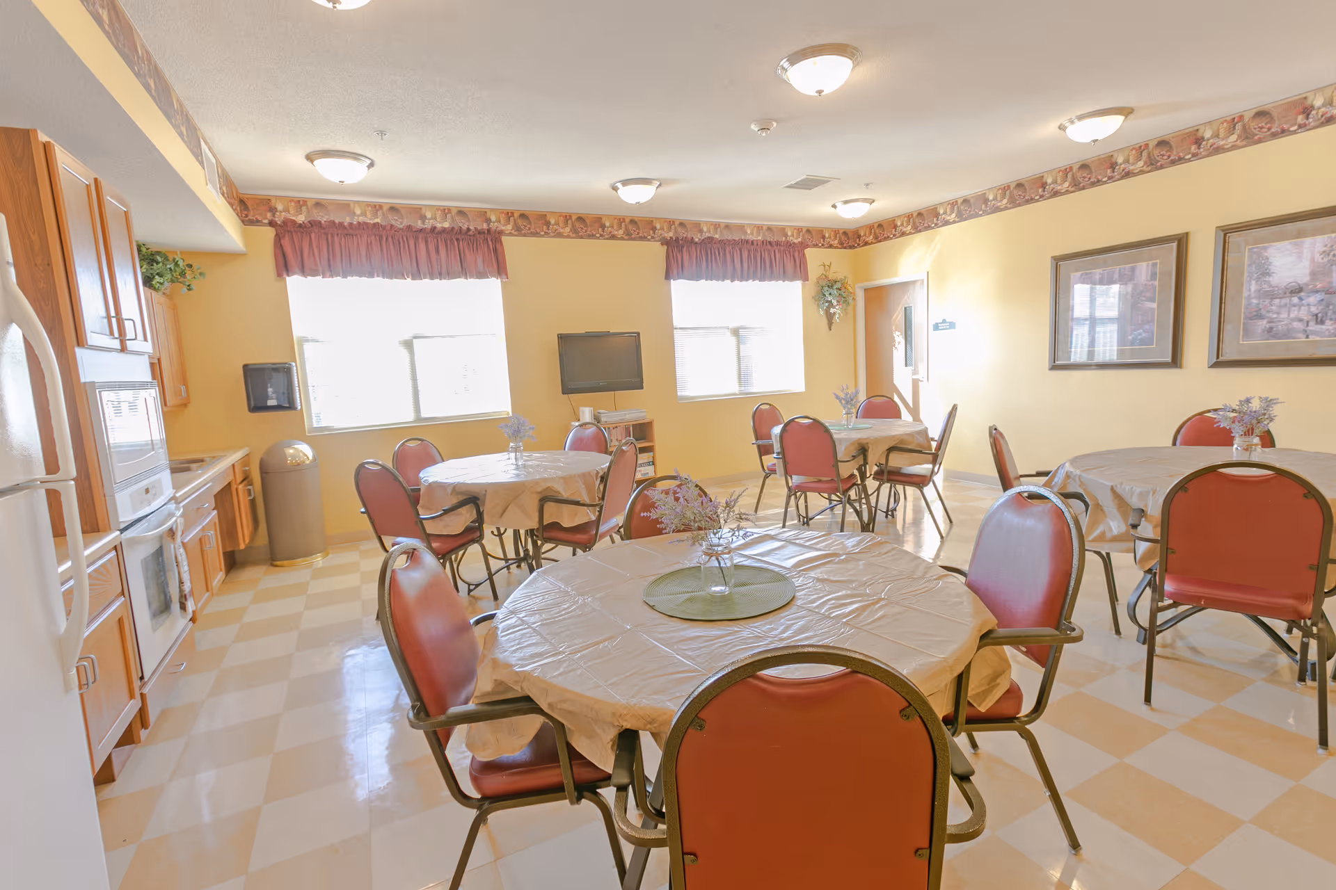 A bright dining room with several round tables covered with beige tablecloths and small floral centerpieces. Each table is surrounded by red cushioned chairs with metal frames. The room has yellow walls with a decorative border near the ceiling, two large windows with red valances, framed artwork on the walls, and ceiling lights. A kitchen area with wooden cabinets, a microwave, and a refrigerator is visible on the left side. A flat-screen TV is mounted on a stand near the windows.