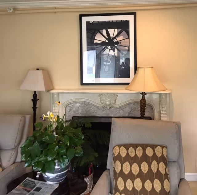 A cozy living room area with two beige armchairs, one featuring a cushion with a leaf pattern. Between the chairs is a small table with a green potted plant and a newspaper. Behind the chairs is a decorative fireplace with a framed black and white photo of a clock above it. Two lamps with beige shades flank the fireplace.