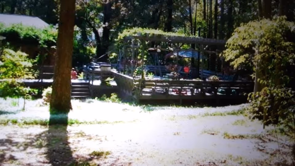 Outdoor area of a senior living facility with a wooden deck surrounded by trees and greenery. The deck has seating and tables under a pergola structure, with sunlight filtering through the trees.