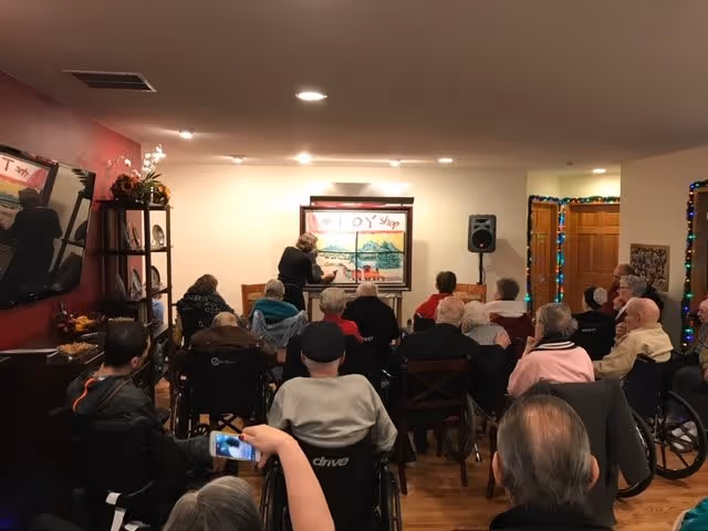 A group of elderly people seated in a room, some in wheelchairs, watching a person at the front who appears to be drawing or writing on a large board labeled 'Candy Shop'. The room has warm lighting, a TV mounted on the wall, and festive string lights around a doorway.