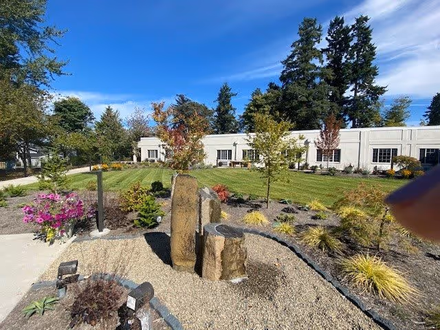 Outdoor garden area with a stone water feature surrounded by gravel and various plants, trees, and flowers. In the background, there is a single-story white building with multiple windows under a clear blue sky.