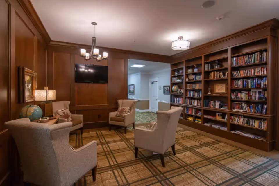 A cozy interior room with wood-paneled walls and a large built-in bookshelf filled with books and decorative items. Four beige upholstered armchairs are arranged around a small wooden table with a globe and lamp. A flat-screen TV is mounted on the wood-paneled wall. The room has a patterned carpet and is softly lit by ceiling lights and a chandelier.