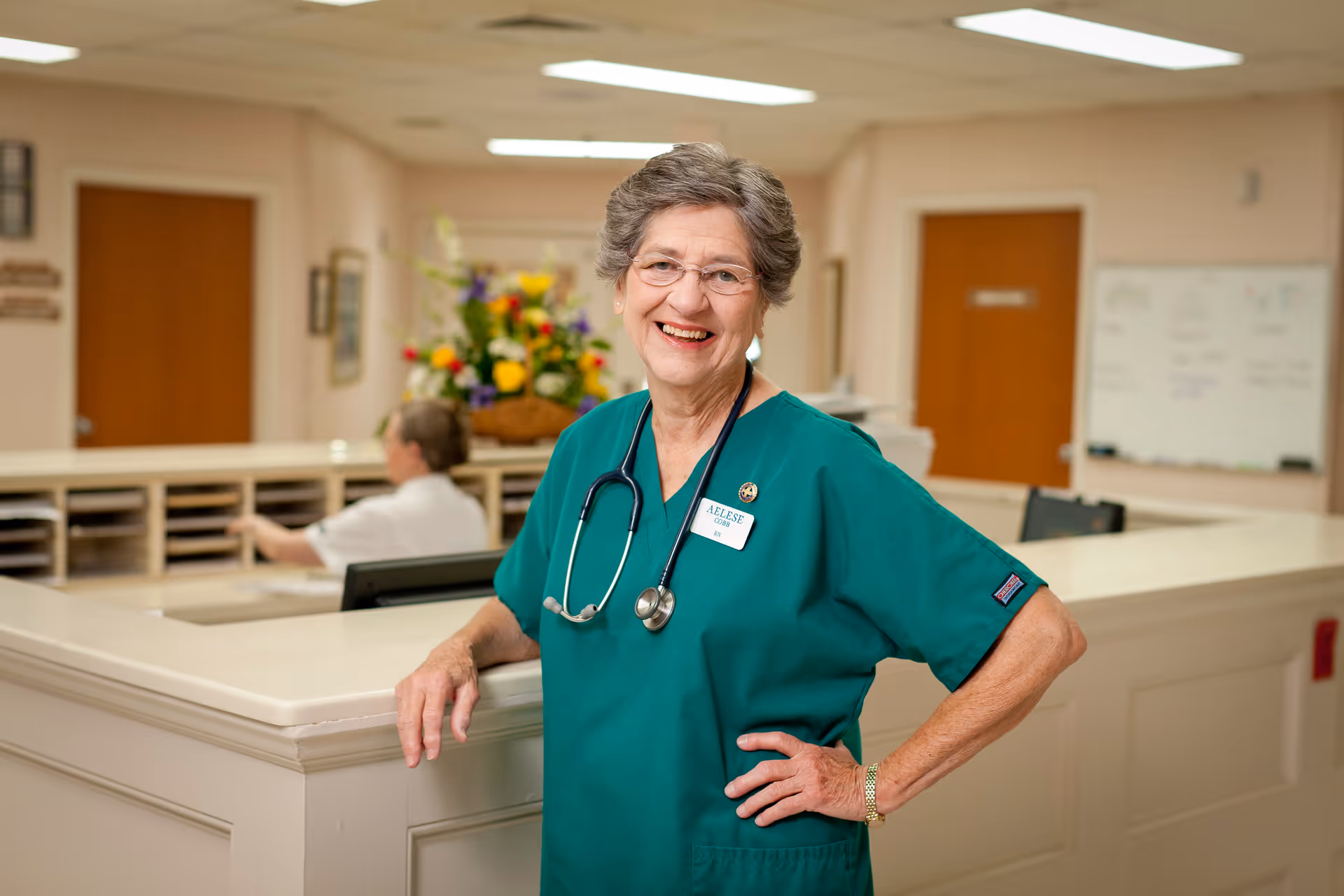 A smiling female nurse in green scrubs with a stethoscope around her neck stands confidently in a healthcare facility reception area. Behind her, another staff member is seated at a desk with a computer and mail slots, and a bouquet of colorful flowers is visible in the background.