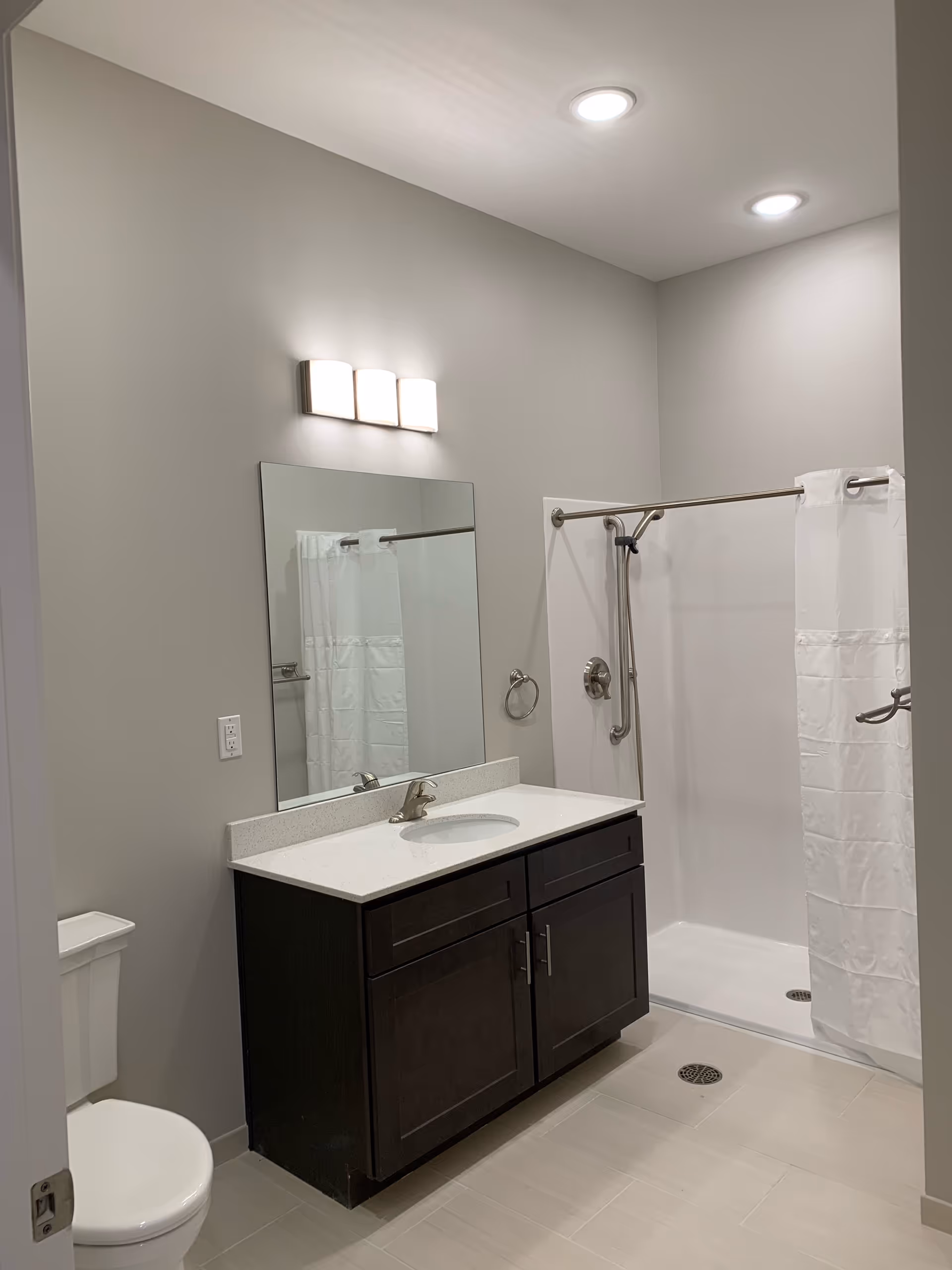 A modern bathroom with a white toilet, a dark wood vanity with a white countertop and sink, a large rectangular mirror above the sink, a three-light fixture above the mirror, and a walk-in shower with a white curtain and stainless steel grab bars.