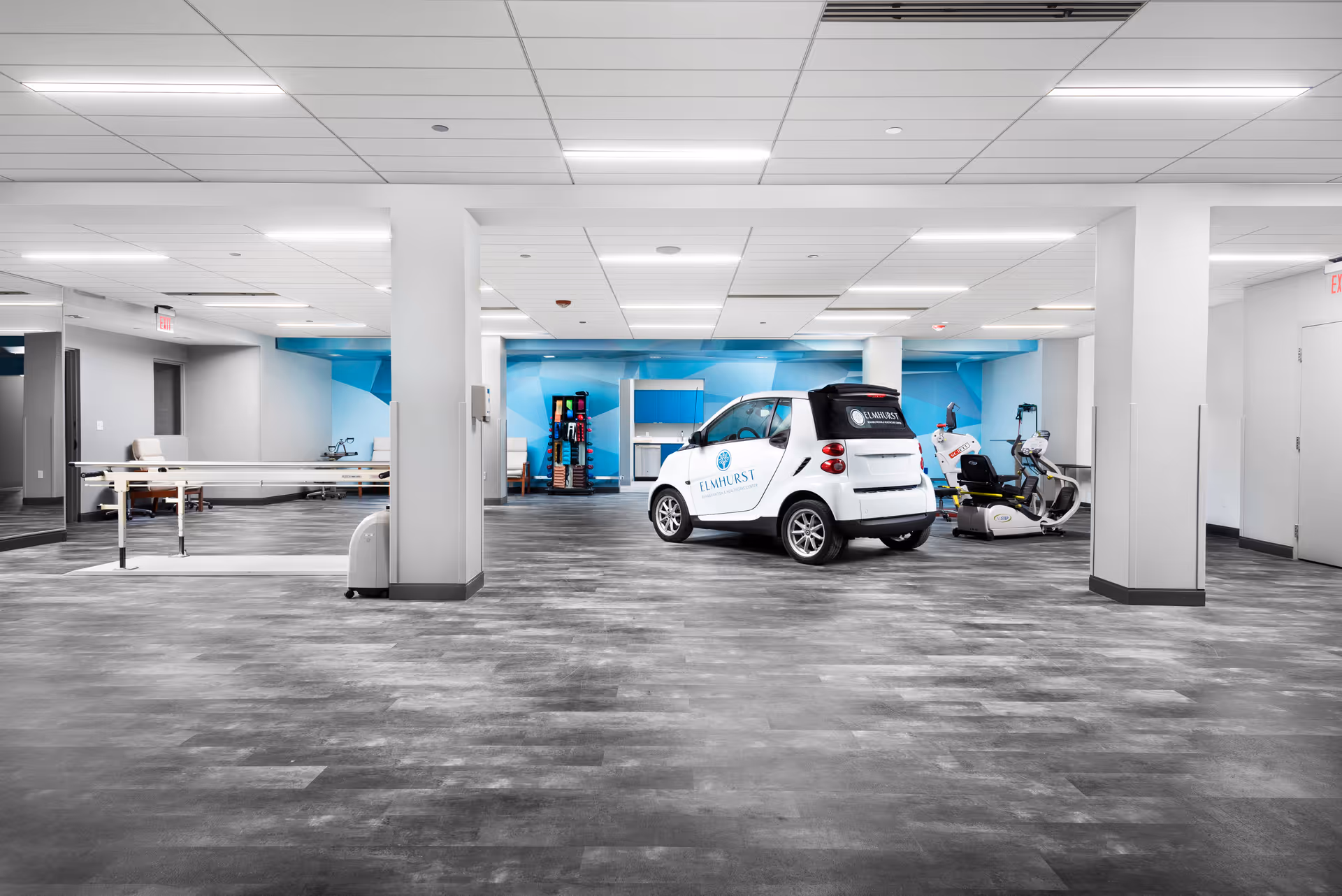A spacious rehabilitation therapy room with exercise equipment, parallel bars, and a small Elmhurst-branded car parked inside.