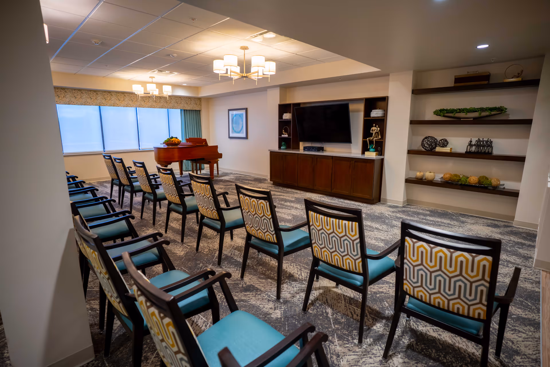 A community activity room with rows of patterned chairs facing a wall-mounted TV, a grand piano, and built-in shelving.