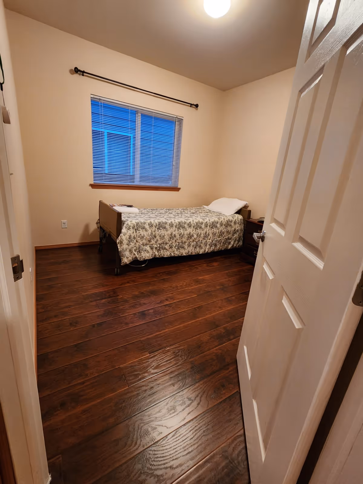 A small bedroom with a single bed covered in a floral patterned blanket and a white pillow. The room has wooden flooring, beige walls, a window with closed blinds, and a nightstand next to the bed. The door to the room is partially open.