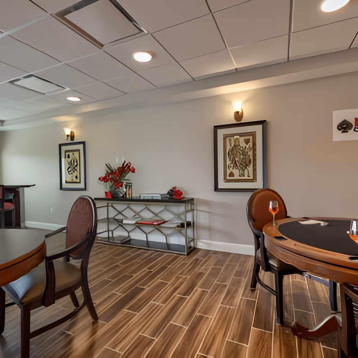 Interior room with wooden flooring, featuring a round table with chairs around it, a console table against the wall with decorative items and red flowers, two framed artworks on the wall, and wall-mounted lights above each artwork.