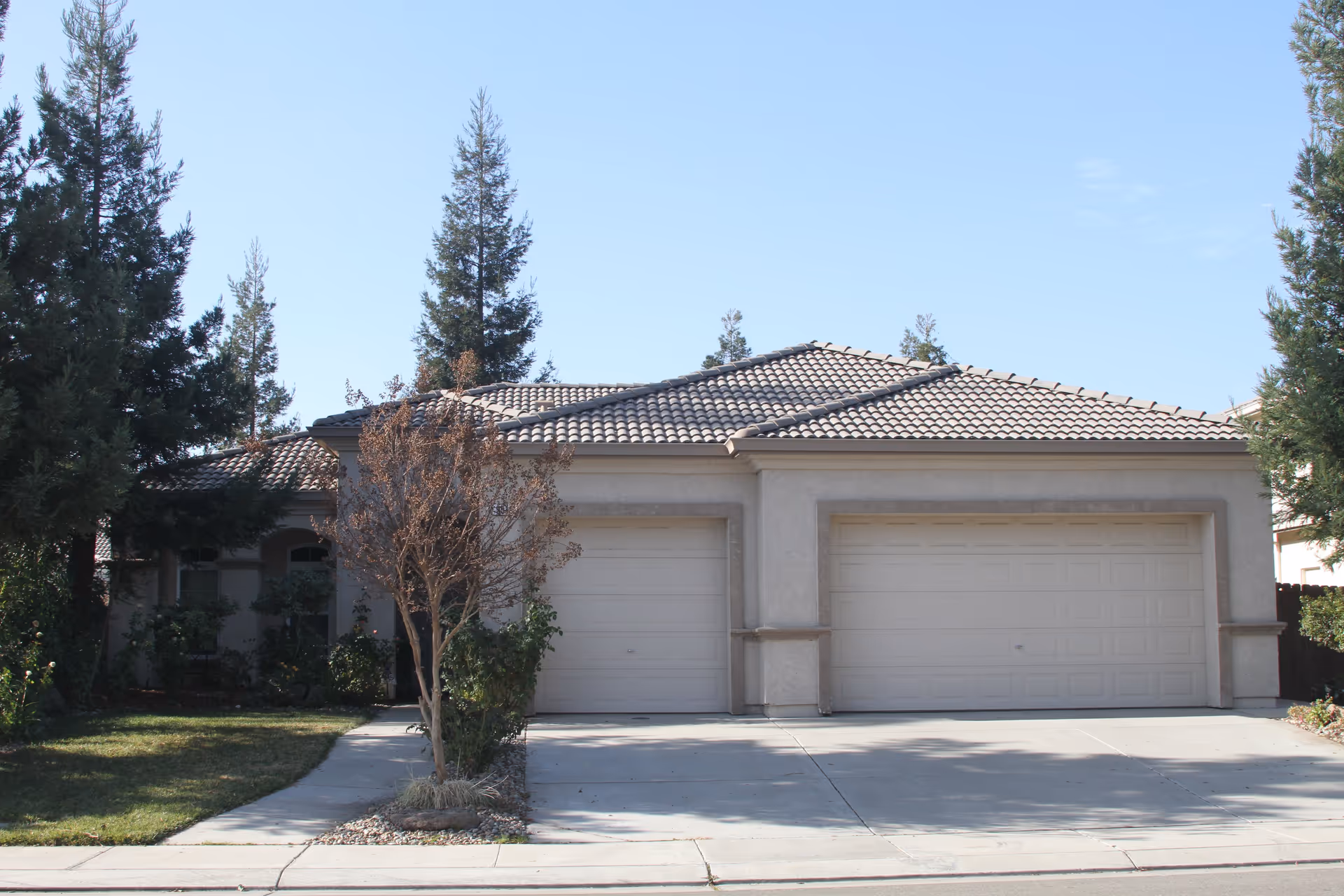 Front exterior view of a single-story house with a tiled roof, two garage doors, a driveway, and trees on either side.