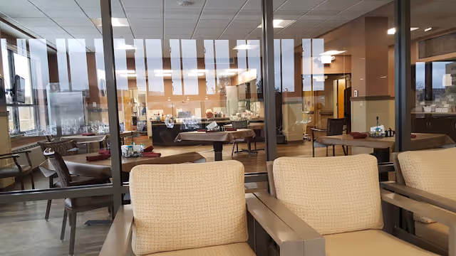 Interior view of a senior living facility dining area with tables and chairs arranged neatly. The foreground shows cushioned armchairs separated by a small table. Large windows and glass partitions allow natural light to fill the space, revealing a clean and organized dining room setup with place settings and napkins on the tables.