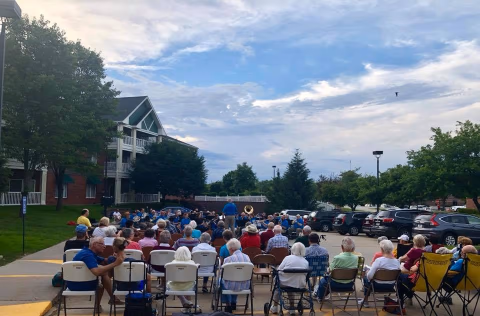 A group of elderly people seated outdoors in folding chairs watching a band perform in a parking lot area near a residential building under a partly cloudy sky.