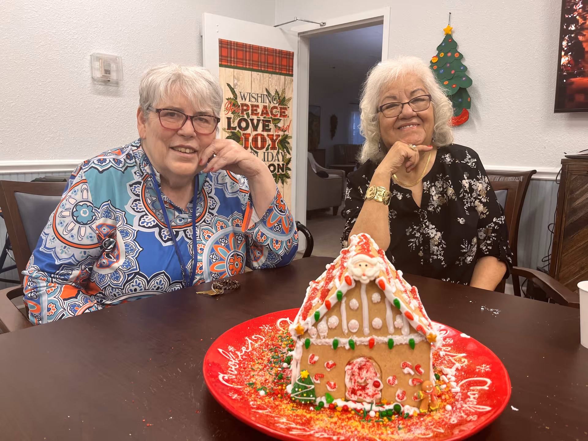 Two elderly women sitting at a table smiling with a decorated gingerbread house on a red plate in front of them. Behind them is a door with a holiday-themed hanging that reads 'Wishing you peace love joy this holiday season' and a small Christmas tree decoration on the wall.