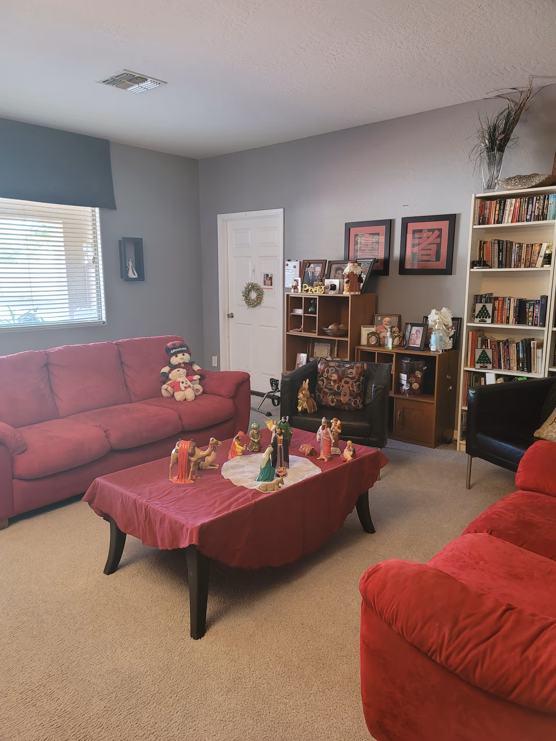 A cozy living room with two red sofas and a black armchair arranged around a coffee table covered with a red cloth and decorated with a nativity scene. The room has beige carpet, a window with blinds and a blue valance, a white door with a small wreath, a wooden shelving unit with framed photos and decorative items, and a tall white bookshelf filled with books and holiday decorations.