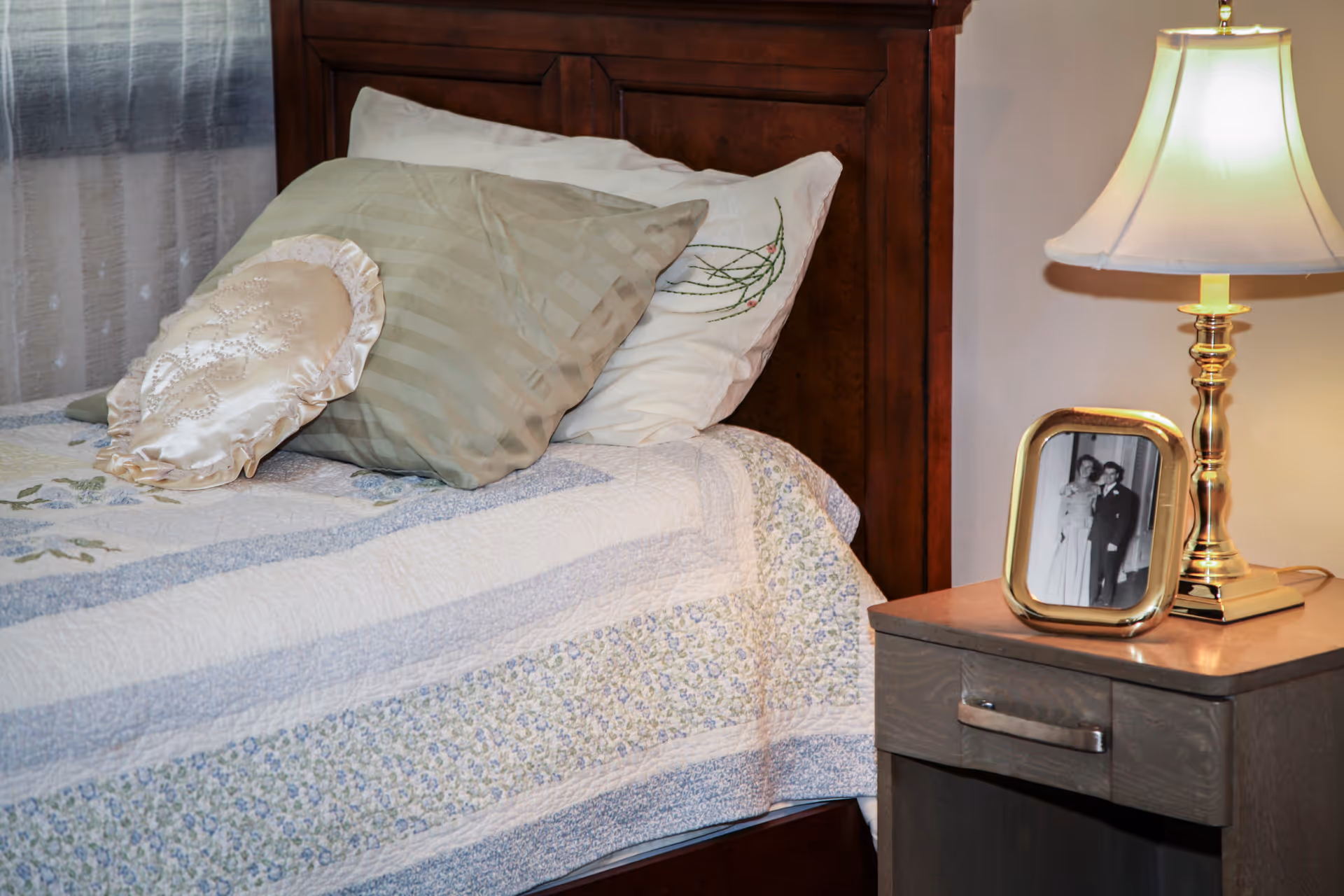 A neatly made bed with a wooden headboard, adorned with three pillows including a decorative round pillow. Next to the bed is a wooden nightstand with a brass table lamp and a framed black-and-white photograph of a couple.